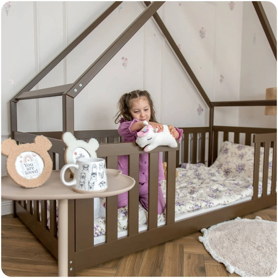 A girl is playing with a plush toy by the safety rail of a dark chocolate-coloured house-shaped bed. The room has a feminine design with purple accents and floral motifs.