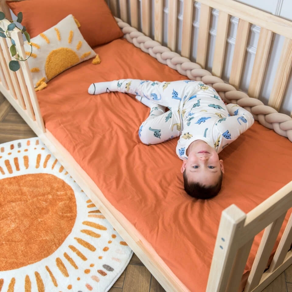 A boy is lying on a pine floor bed with high safety rails. Orange tones dominate the interior, complemented by boho decorations with a sun motif.