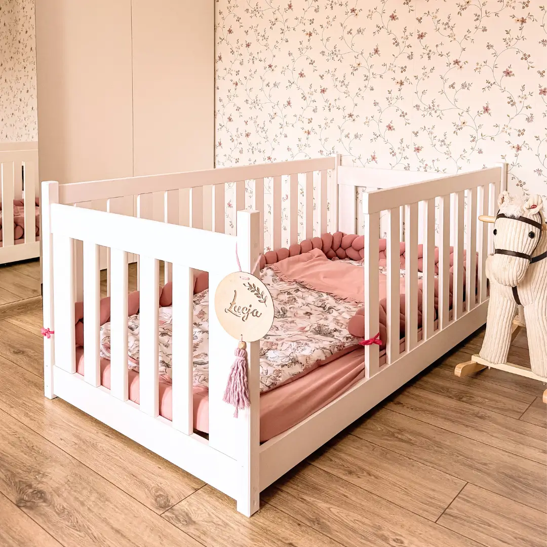 Girl's room. CONCEPT montessori children's bed with high railings. The bed has pink bedding and a protective braid to prevent the child from hitting themselves. Floral wallpaper in the background.