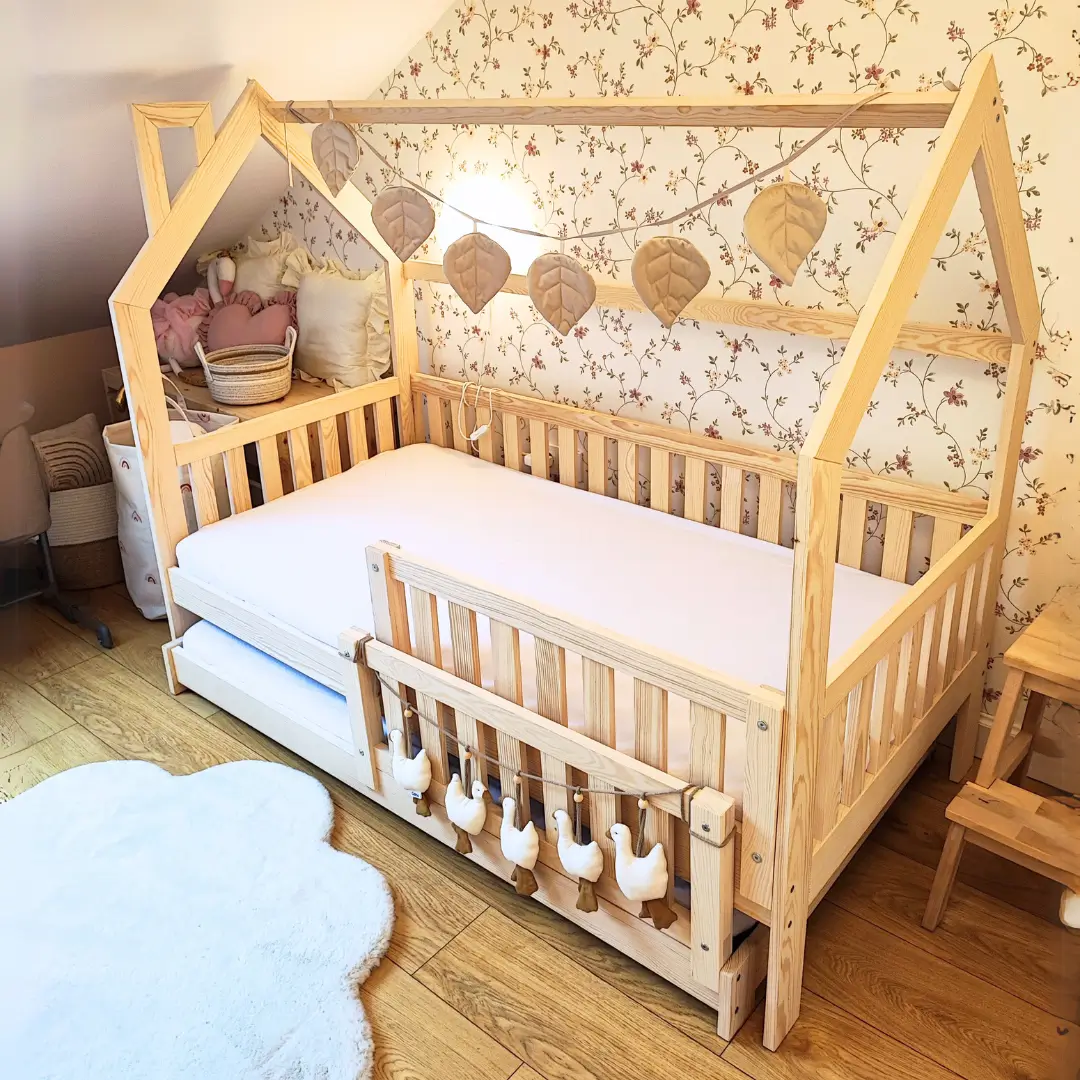 Wooden MONKEY children's bed in the shape of a house with a pull-out bottom bed with a protective railing. Decorated with a garland of leaves and a pendant with geese. In the background, there is floral wallpaper in muted colors.