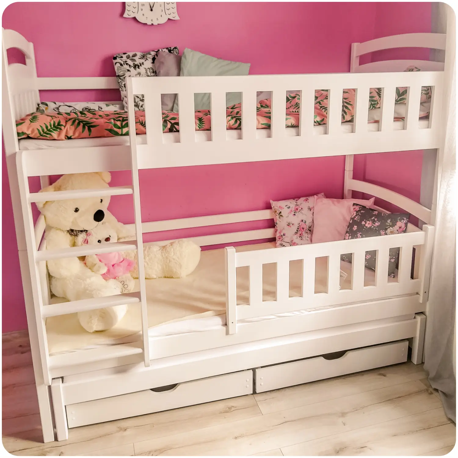 White children's bunk bed BLANKA. A three-person bed with a pull-out bottom sleeping deck. Equipped with high protective railings on the upper and lower levels, a side ladder, and two pull-out drawers. The bed is in a room with pink walls and a light floor. Next to it is a light-transmitting curtain and decorative pillows.