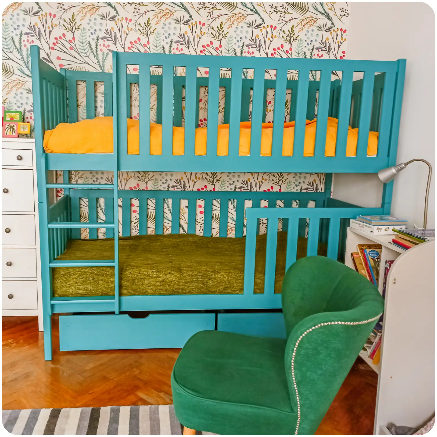 Turquoise children's bunk bed DAWID. It has high protective railings, a vertical ladder, and two pull-out drawers under the bottom bed. Placed against a wall with floral wallpaper. The room has an upholstered armchair, a desk lamp, and a bookshelf with books.