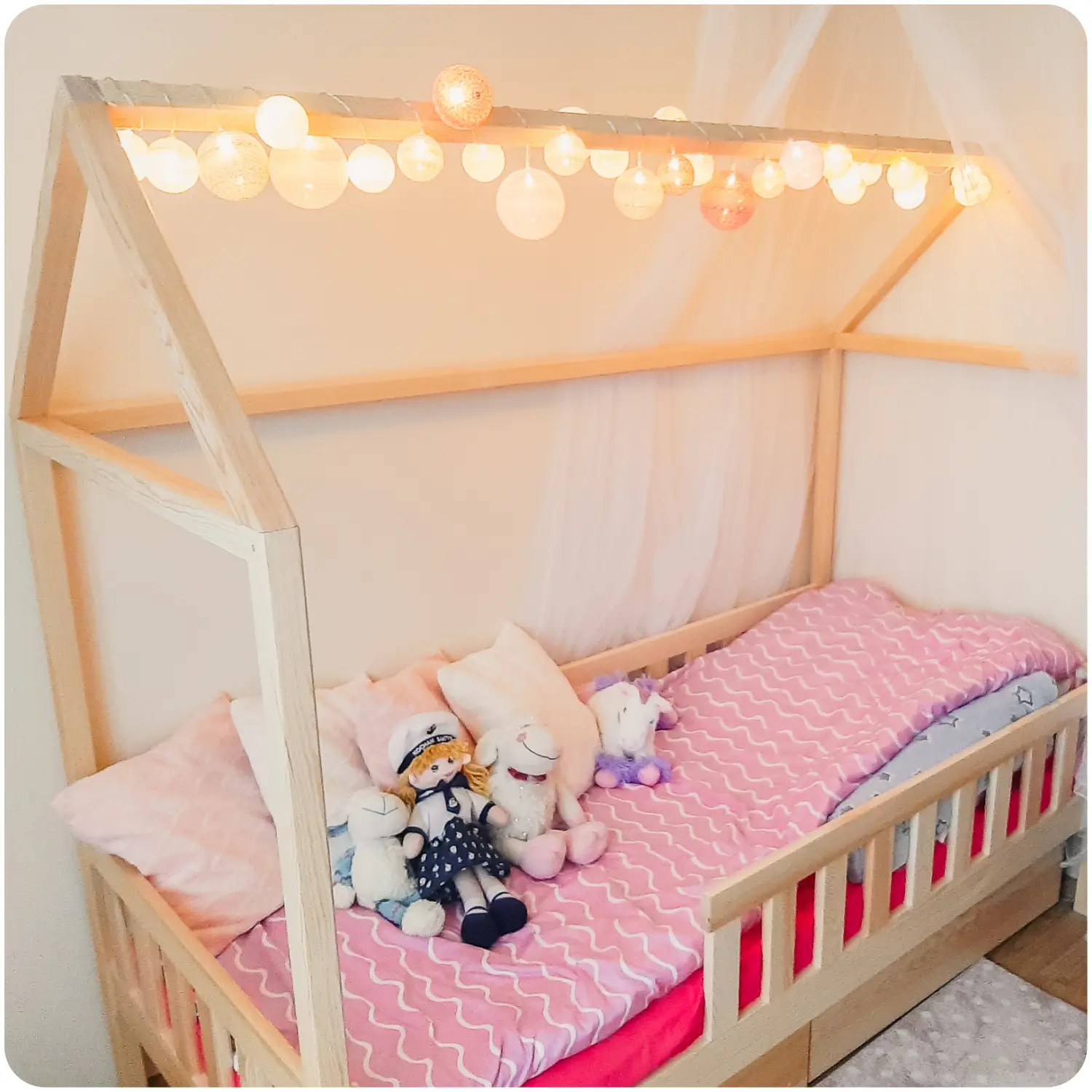 Pine children's bed DOMI in the shape of a house with pull-out drawers, decorated with a white canopy and lights. It has protective railings. The bed has pink bedding, pillows, and stuffed animals.