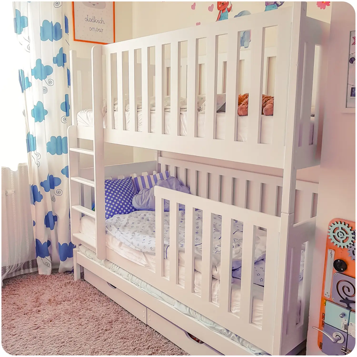White triple bunk bed for children EMILKA with high railings, a pull-out bottom bed, and drawers for bedding or toys. In front of the bed is a pink carpet, and on the wall hangs a curtain with blue clouds.