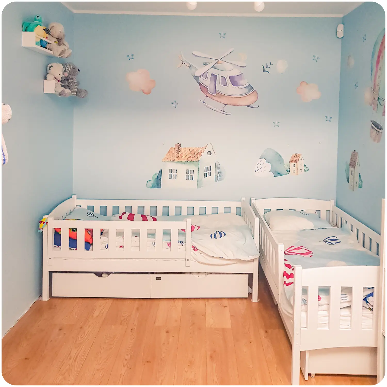 Two white GUCIO children's beds placed in the corner of the room, both with a protective railing and a drawer for bedding. The wall is blue with paintings of a helicopter and houses. The bedding is blue with hot air balloons.