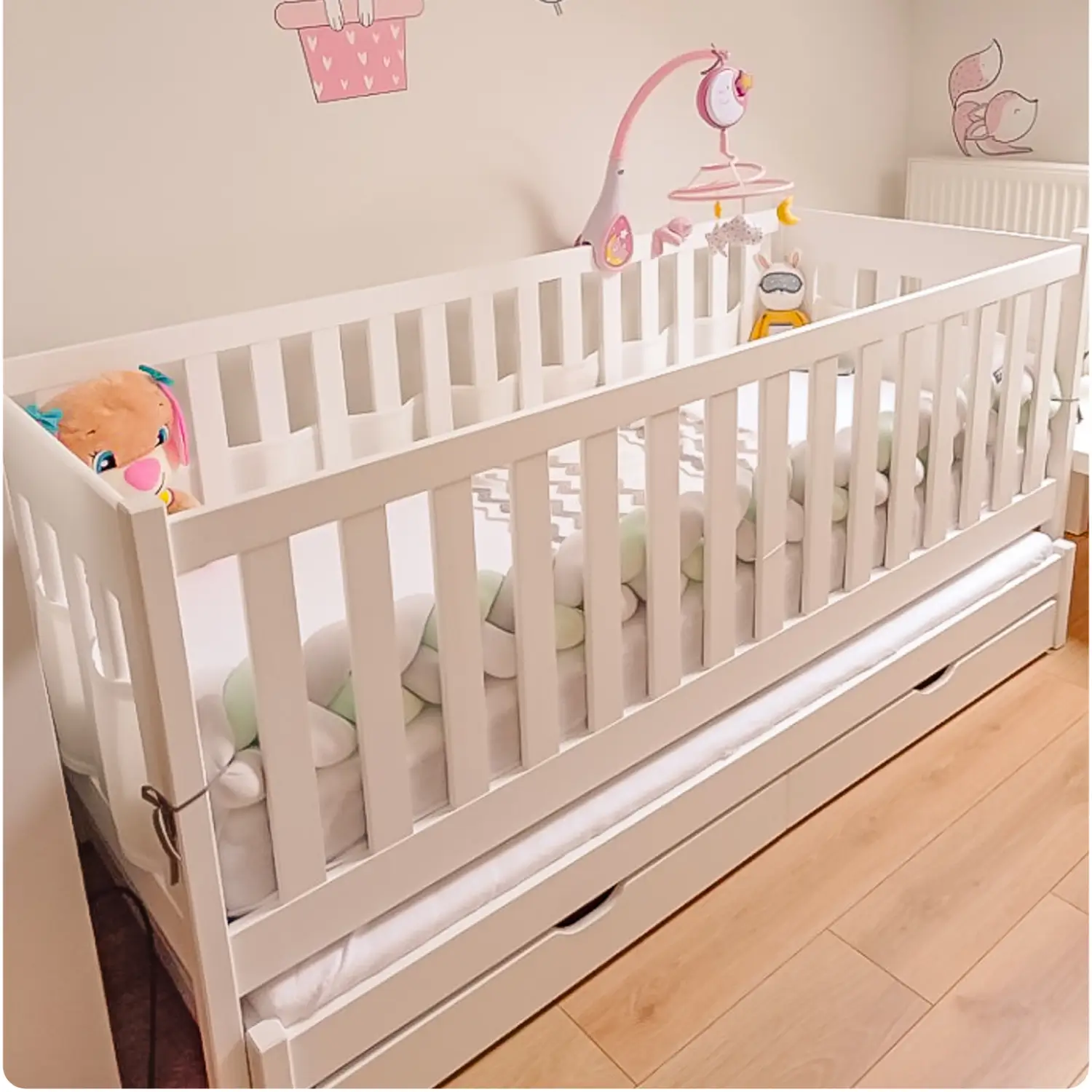 White pine children's bed KAJA with high, fully enclosed railings, with a pull-out second bed and drawers at the bottom. Inside the bed, there is a soft braid bumper and a mobile with toys.