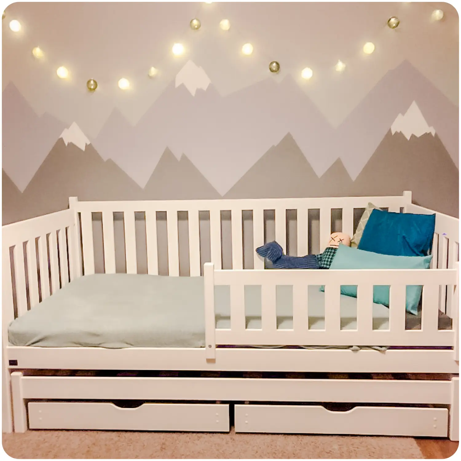 Low double children's bed KAJA with protective railings and a pull-out bed. The model has two spacious drawers. A garland of lights hangs above the bed. The wallpaper has a mountain pattern in muted colors.