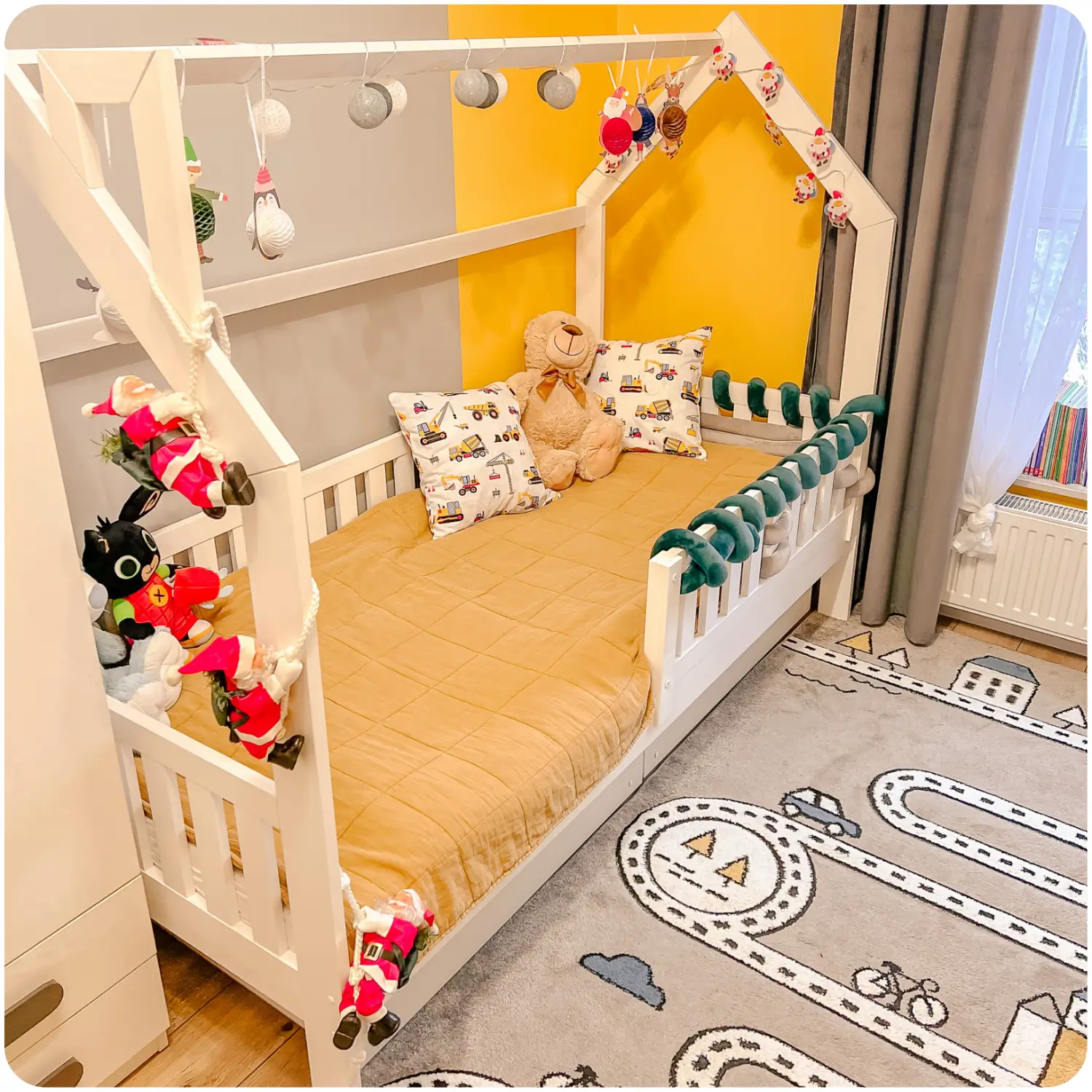 White children's bed NATAN in the form of a house with high protective railings. It is located in a room with a yellow and grey wall. The bed is festively decorated with lights.