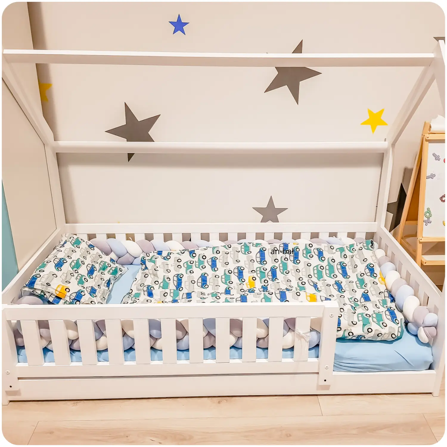 White Montessori children's bed NATAN in the shape of a house, with protective railings and a soft braid bumper. Bedding with colorful cars, and the wall is decorated with stars.