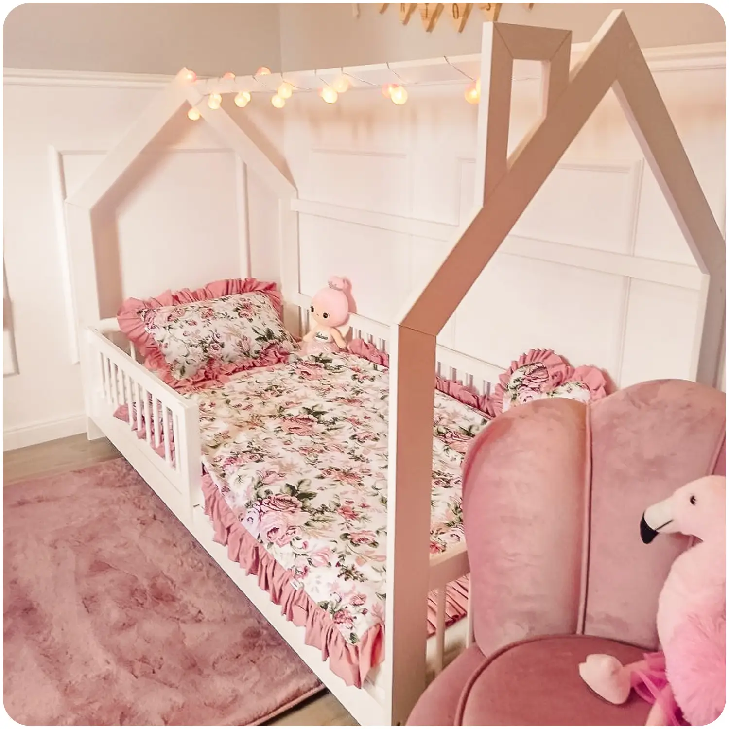 Children's room. White children's bed OSKAR in the shape of a house. The bed is decorated with floral bedding with a pink frill. On the floor is a pink carpet contrasting with the white wall.