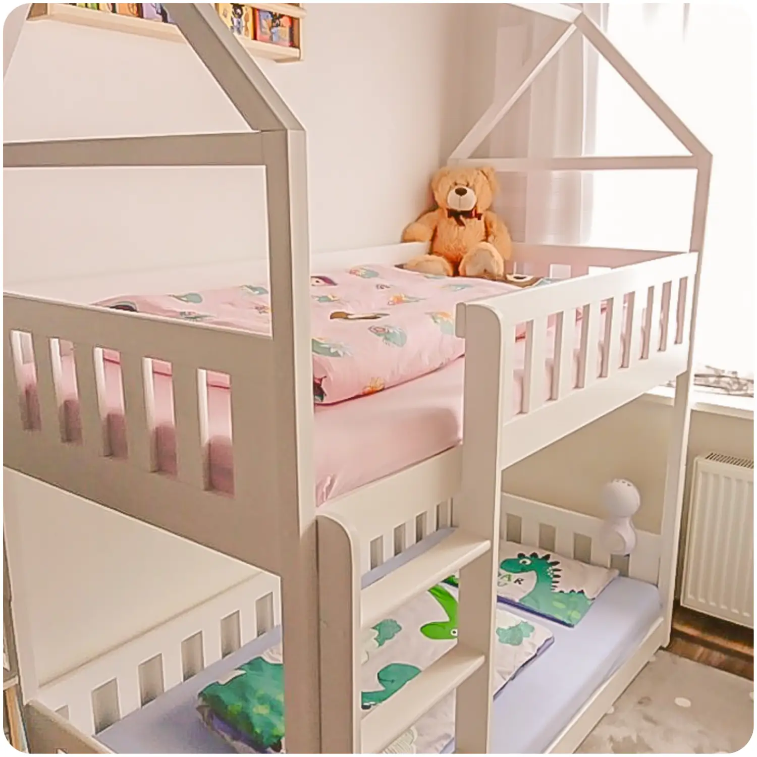 White bunk bed POLA for children in the shape of a house. Equipped with a vertical ladder. Placed against a light wall and a window with a curtain. A teddy bear is on the top bunk.